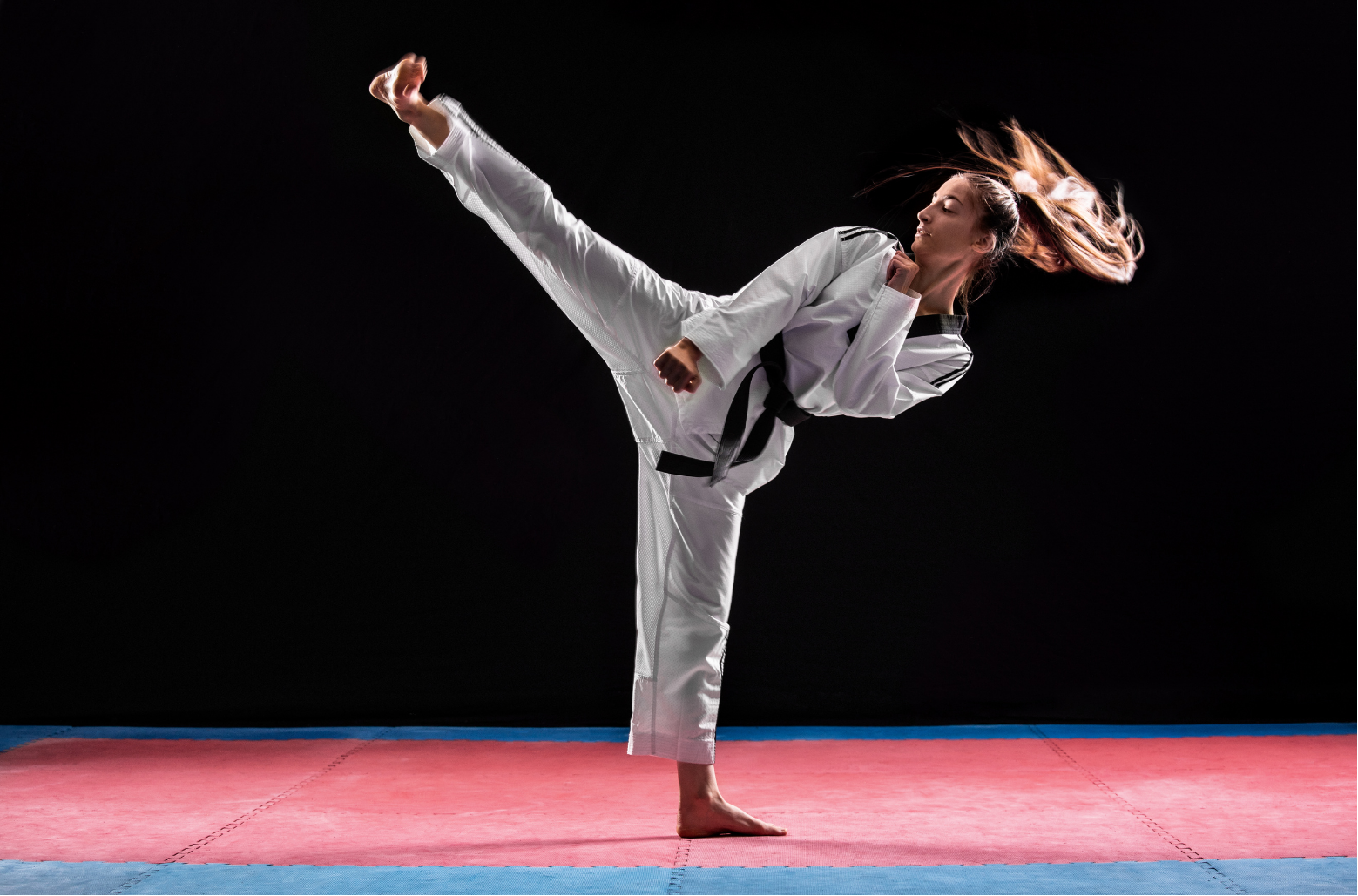 Martial artist in white gi performing a high side kick on a red mat against a black background
