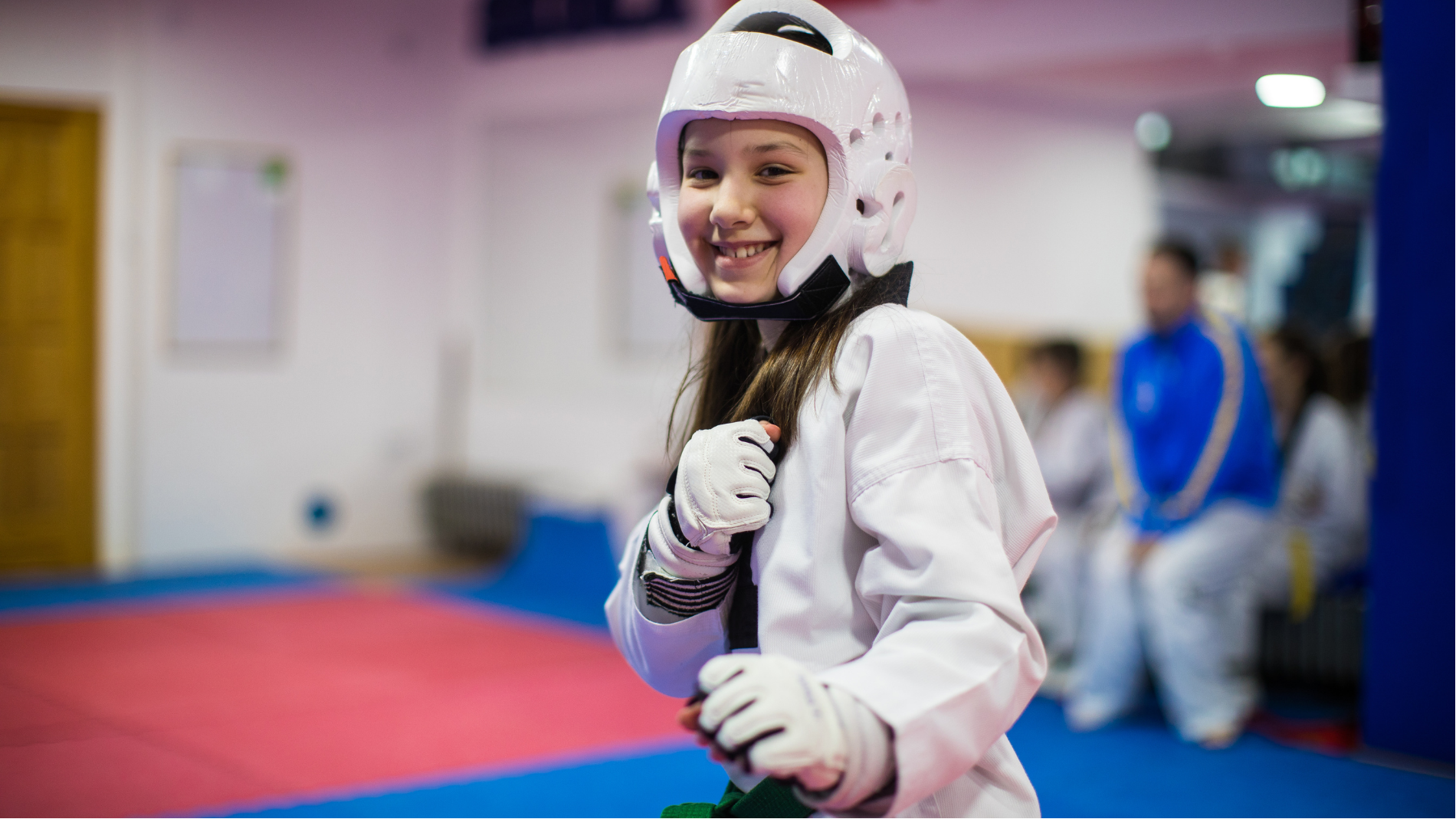 Smiling martial artist in white protective gear and helmet, standing on a mat in a gym.