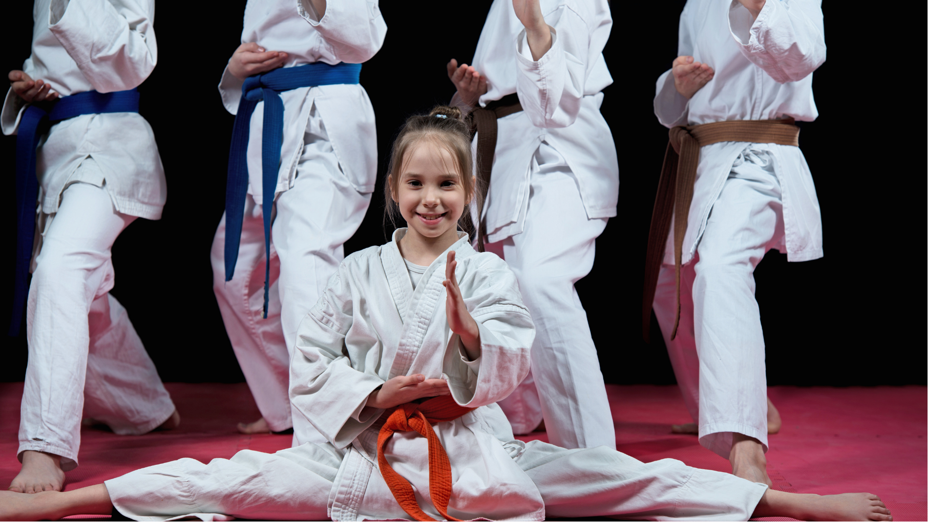 Karate students in white uniforms pose on a red mat, with a child seated front and center holding an orange belt.