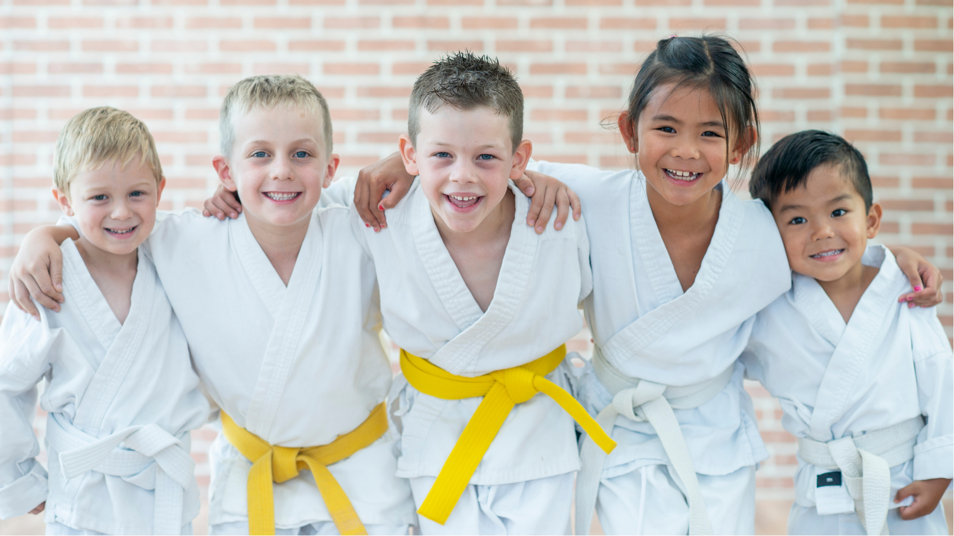 Five children in white karate uniforms standing together, smiling in a bright studio, some with yellow belts