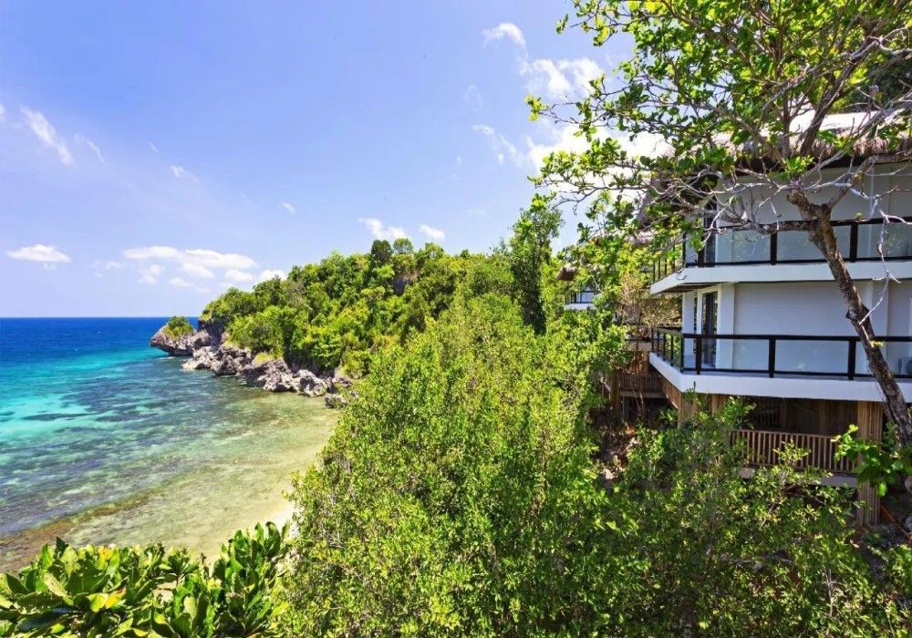 A large white house is sitting on top of a lush green hill overlooking the ocean.