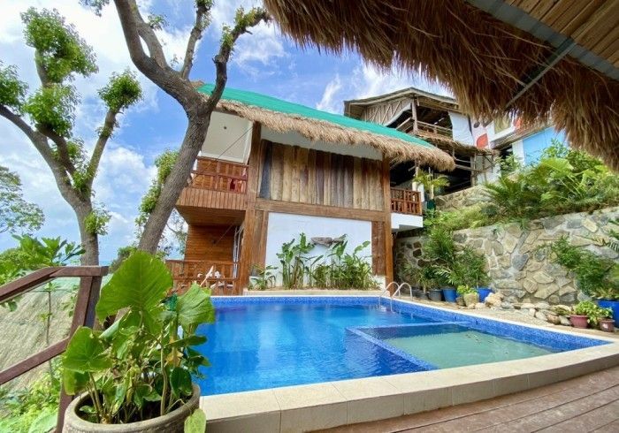 A large swimming pool in front of a house with a thatched roof.