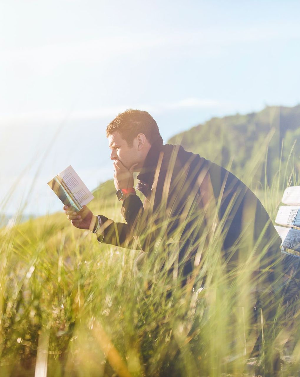 A man is sitting on a bench in the grass reading a book.