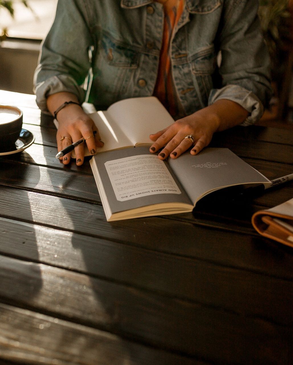 A woman is sitting at a table reading a book and writing in a notebook.