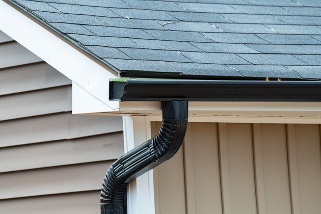 Black rain gutter attached to a house with gray shingles and beige siding.