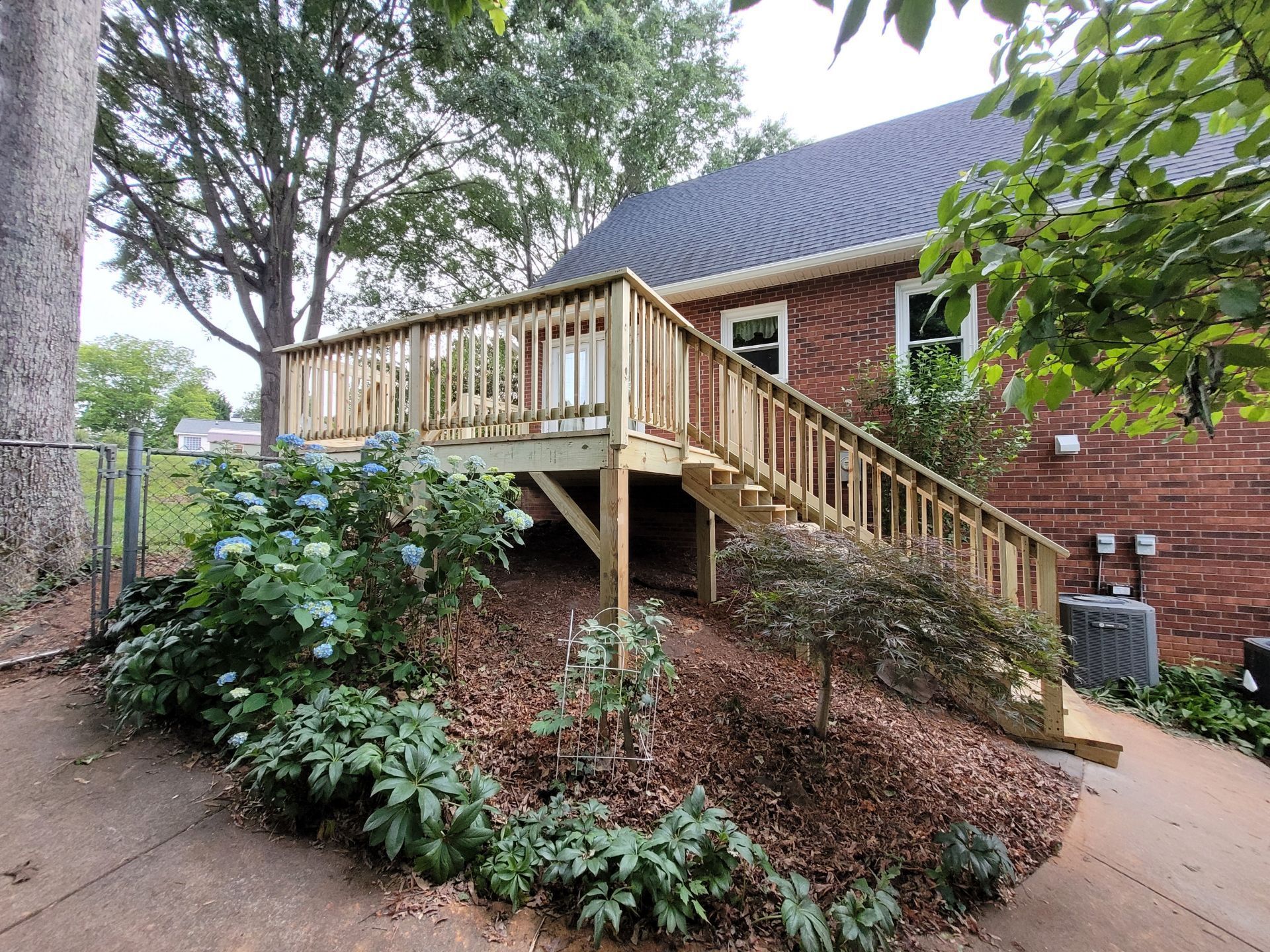 Wooden deck with stairs next to a brick building, set on a sloped, mulched area with greenery.