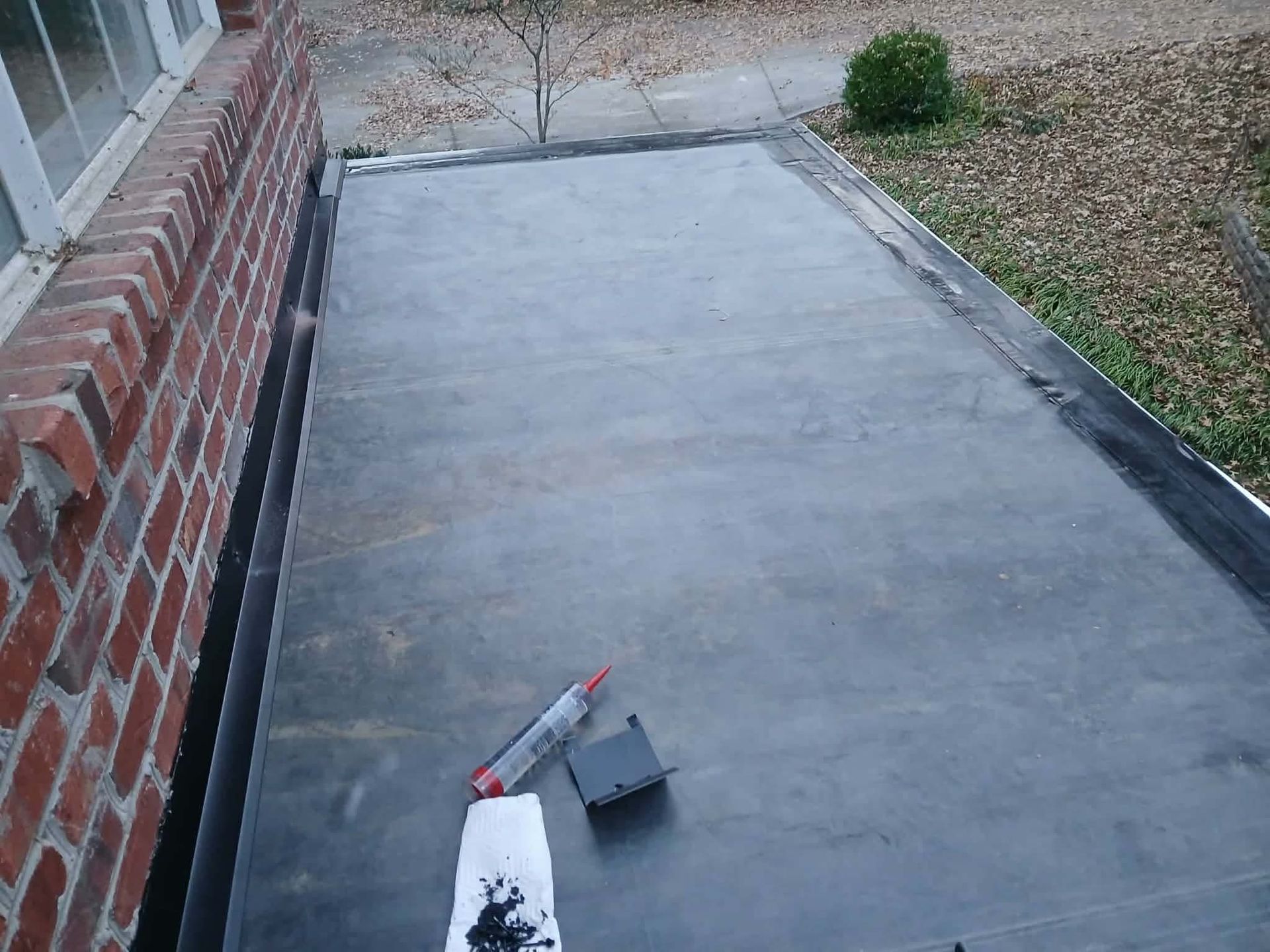 Flat roof with tools, against a brick wall. Black sealant and cartridge sit on the dark surface.