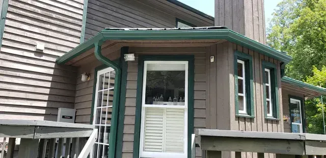 Exterior of a house with brown siding, green trim, and a white window with shutters, on a sunny day.
