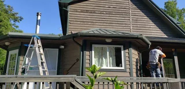 Man on ladder working on a house with a deck. A ladder is leaning against the roof.