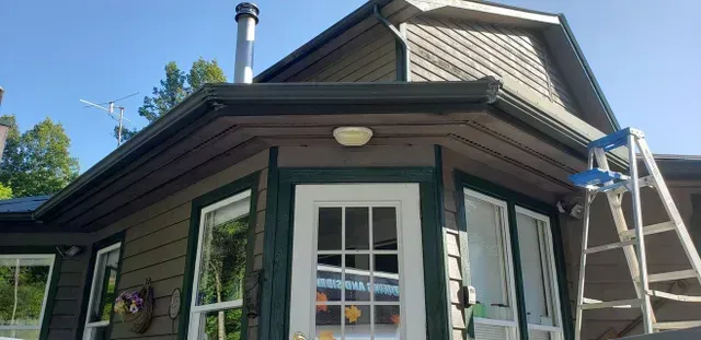 House exterior with green trim, white door, windows. Ladder up against roof, blue sky.