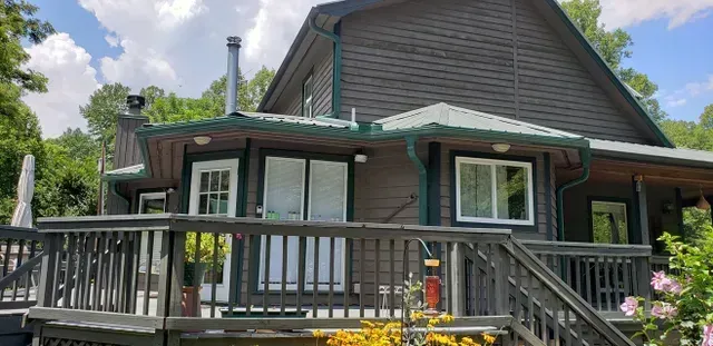 A two-story dark brown house with green trim and a wooden deck. The sky is blue with fluffy white clouds.