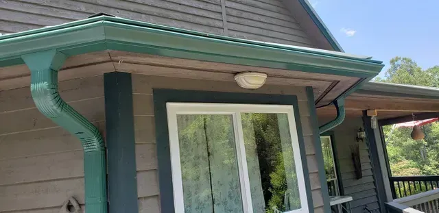 Corner of a house with green gutters and trim, gray siding, and a white window with reflected trees.