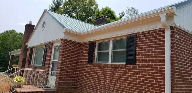 Brick house with white trim, black shutters, and a metal roof. Trees are in the background.