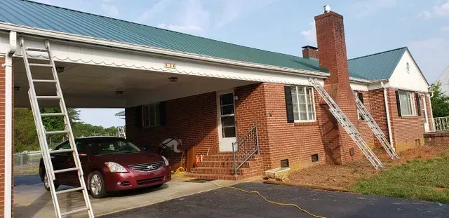 Red brick house with green metal roof, a car parked under a carport, and ladders against the house.