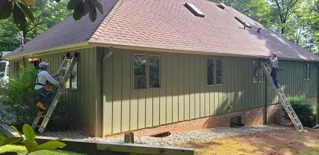 Two workers on ladders are working on the side of a green house with a brown roof.