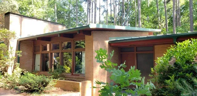 Modern, single-story house with large windows, set in a wooded area. Green roof and brown brick exterior.