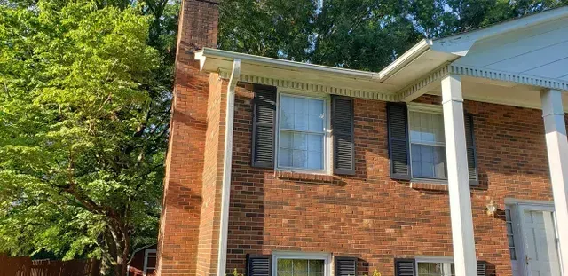 Red brick house with black shutters, white trim, and a porch. Green trees in the background.
