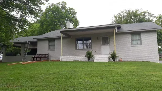 Gray brick house with covered porch, small bushes, and green lawn.