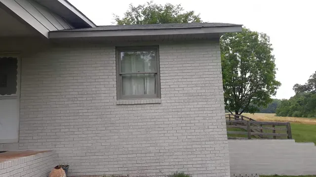 Exterior view of a brick house with a window, painted light gray, with a yard and trees in the background.