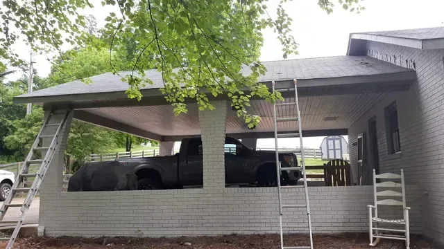 A truck parked under a carport attached to a brick house, ladders and a chair are visible.