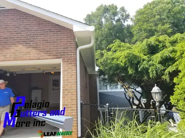 Exterior view of a brick house with white gutters. A person is visible near the open garage. Lush green trees are nearby.