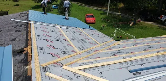 Workers installing a metal roof, with blue panels, on a house. Grass and a red car are visible.