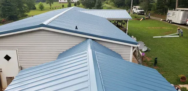 Blue metal roof with a person working on the side of a building, green grass, and trees in the background.