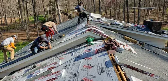 Construction workers installing roofing materials on a roof with a wooded backdrop.