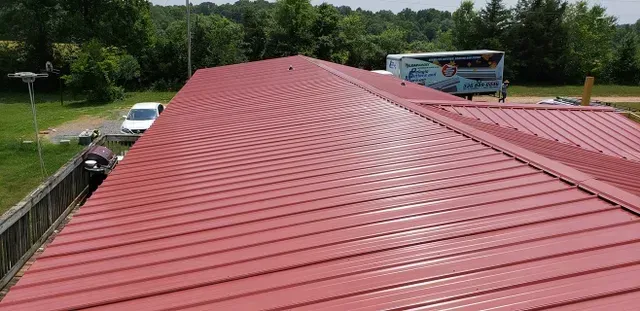 Red metal roof on a building, with a white vehicle and trees in the background.