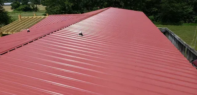 Red metal roof on a building, angled view, showing ridges, gutter, and surrounding trees and field.