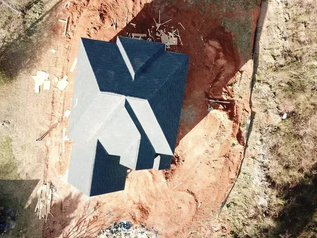 Aerial view of a house under construction with dark gray roof, surrounded by dirt and construction materials.