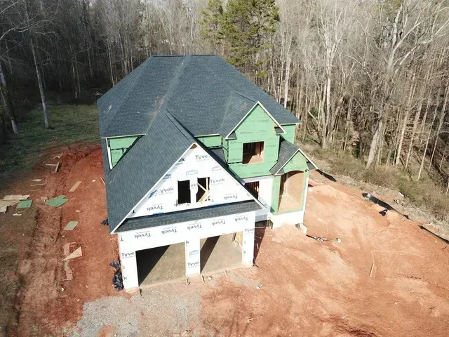 House under construction with dark roof, two-car garage, and surrounding bare trees on a hillside.