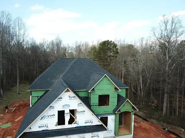 New house under construction, green wrap, black roof, surrounded by trees and red dirt.