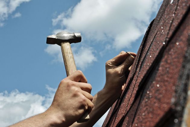 Hands hammering a nail into a roof shingle against a blue sky with clouds.