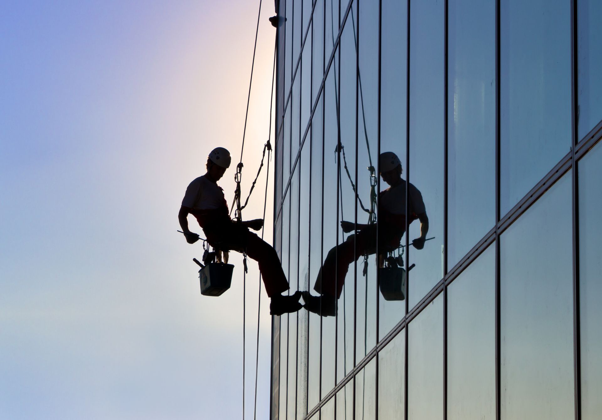 Silhouetted window washers suspended on a glass skyscraper, cleaning windows.