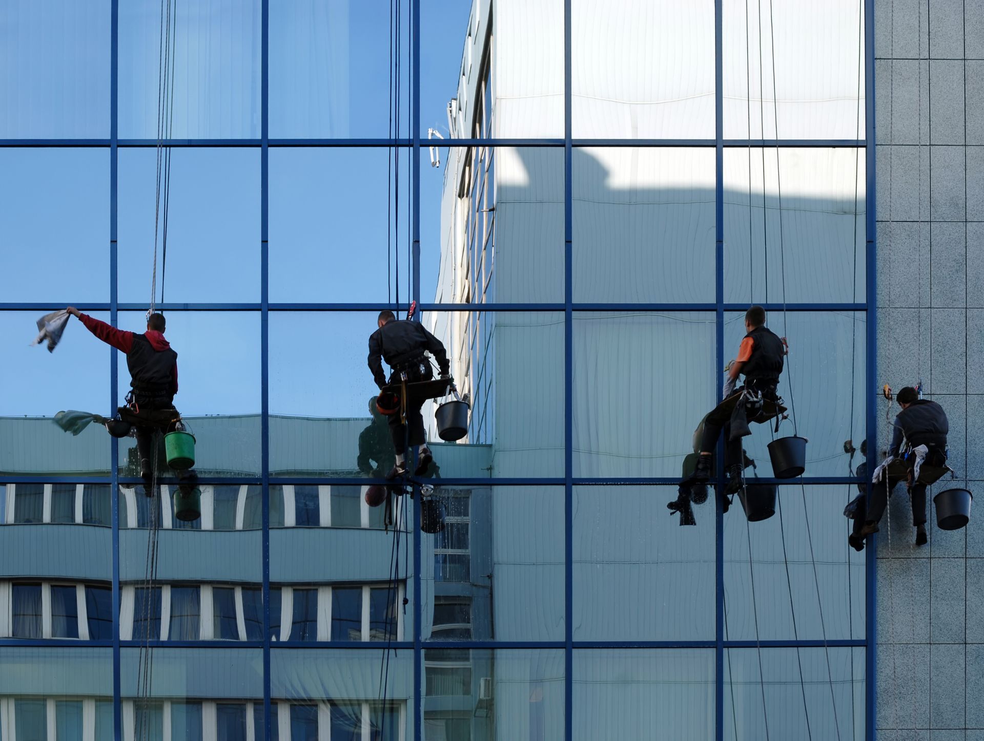 Window washers suspended on a tall building with blue-tinted glass, cleaning exterior windows.