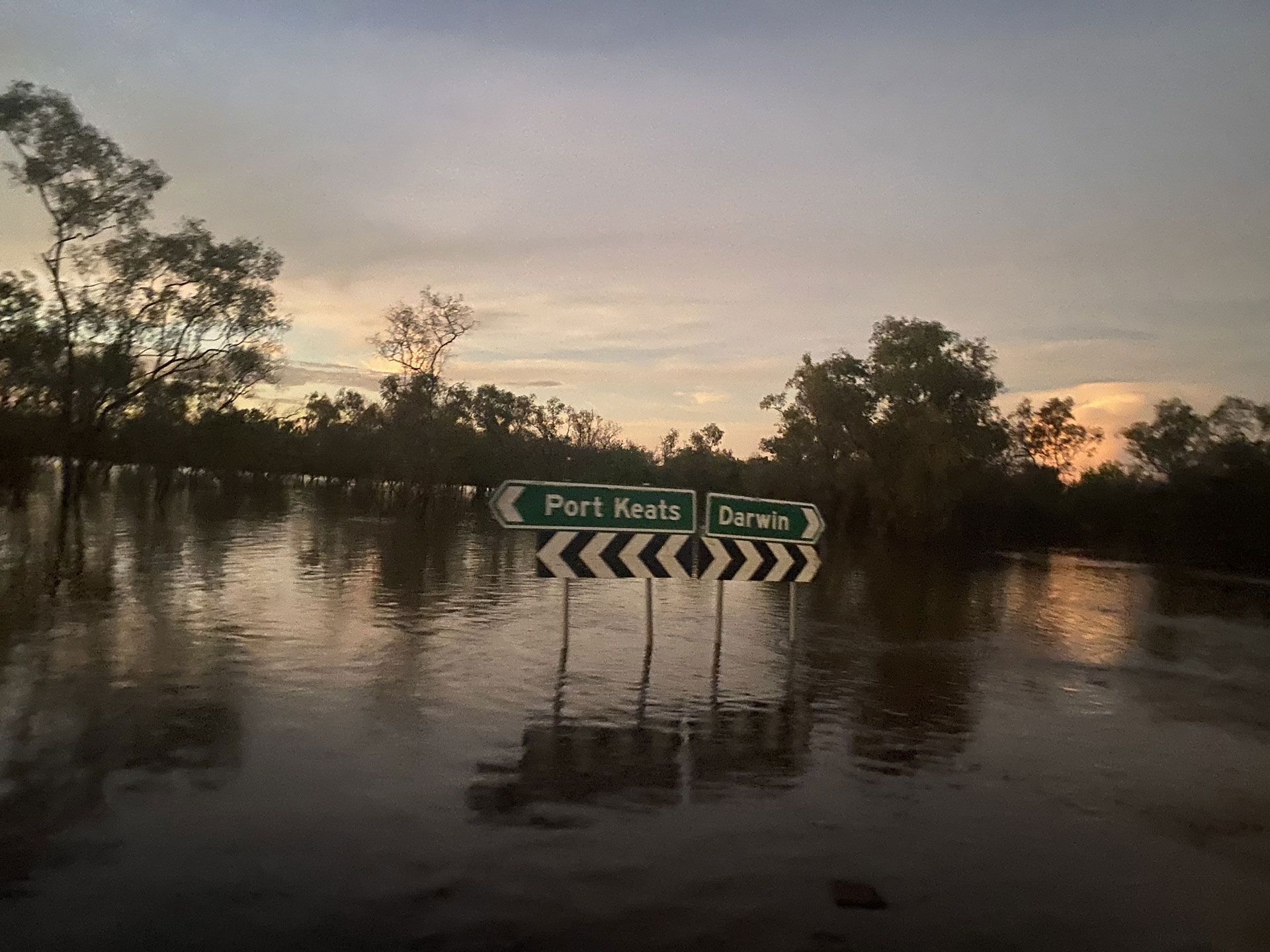 Flooded road with submerged sign pointing to Port Keats and Darwin at sunset.