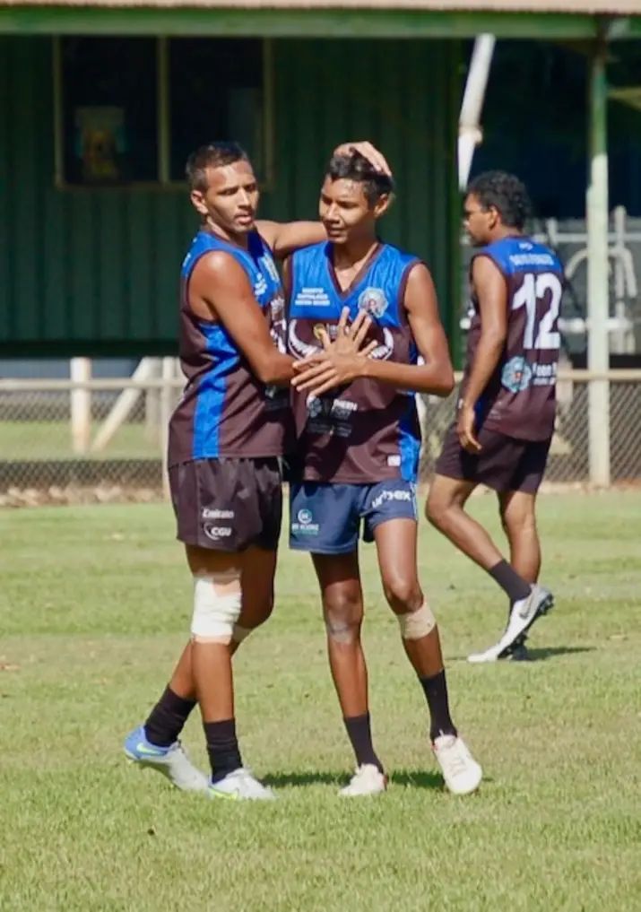 Three young men in athletic gear on a grassy field. Two are embracing; one has hand on the other's chest.