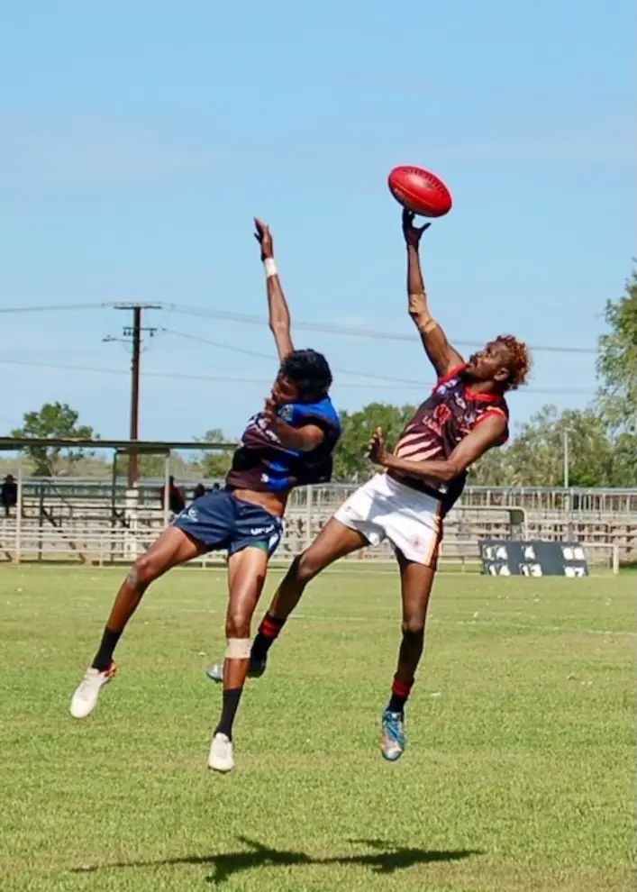 Two Indigenous men leap to catch an oval football on a green field under a blue sky.
