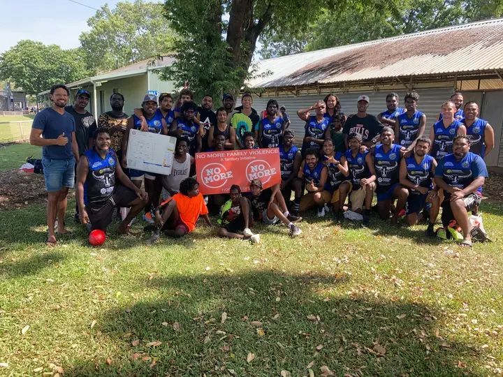 Group of people, mostly in blue team jerseys, pose outdoors with a banner.