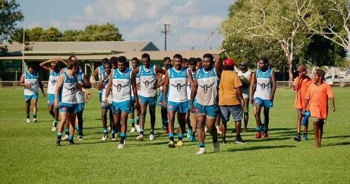 A group of Indigenous Australian men in team uniforms walk on a grassy field; bright sky.