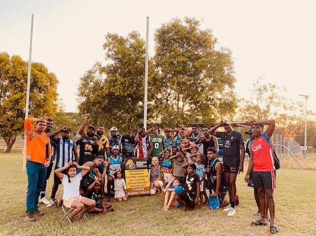 Group of people outdoors, saluting near a sign. Trees and goalposts visible.