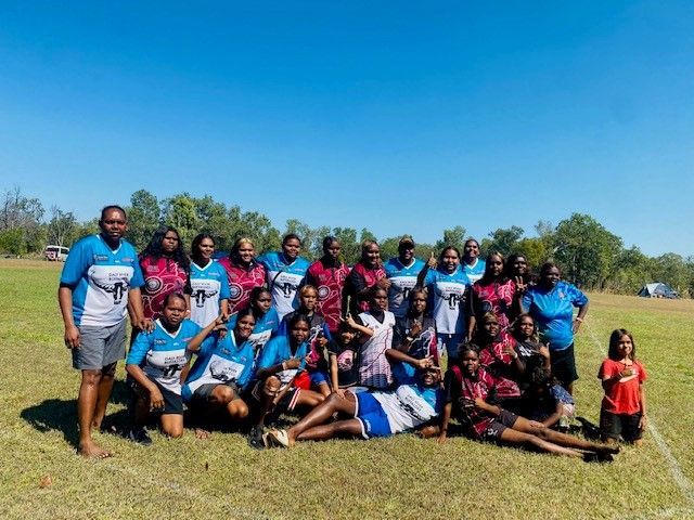 Group of women posing for a photo on a grassy field under a blue sky, wearing sports jerseys.