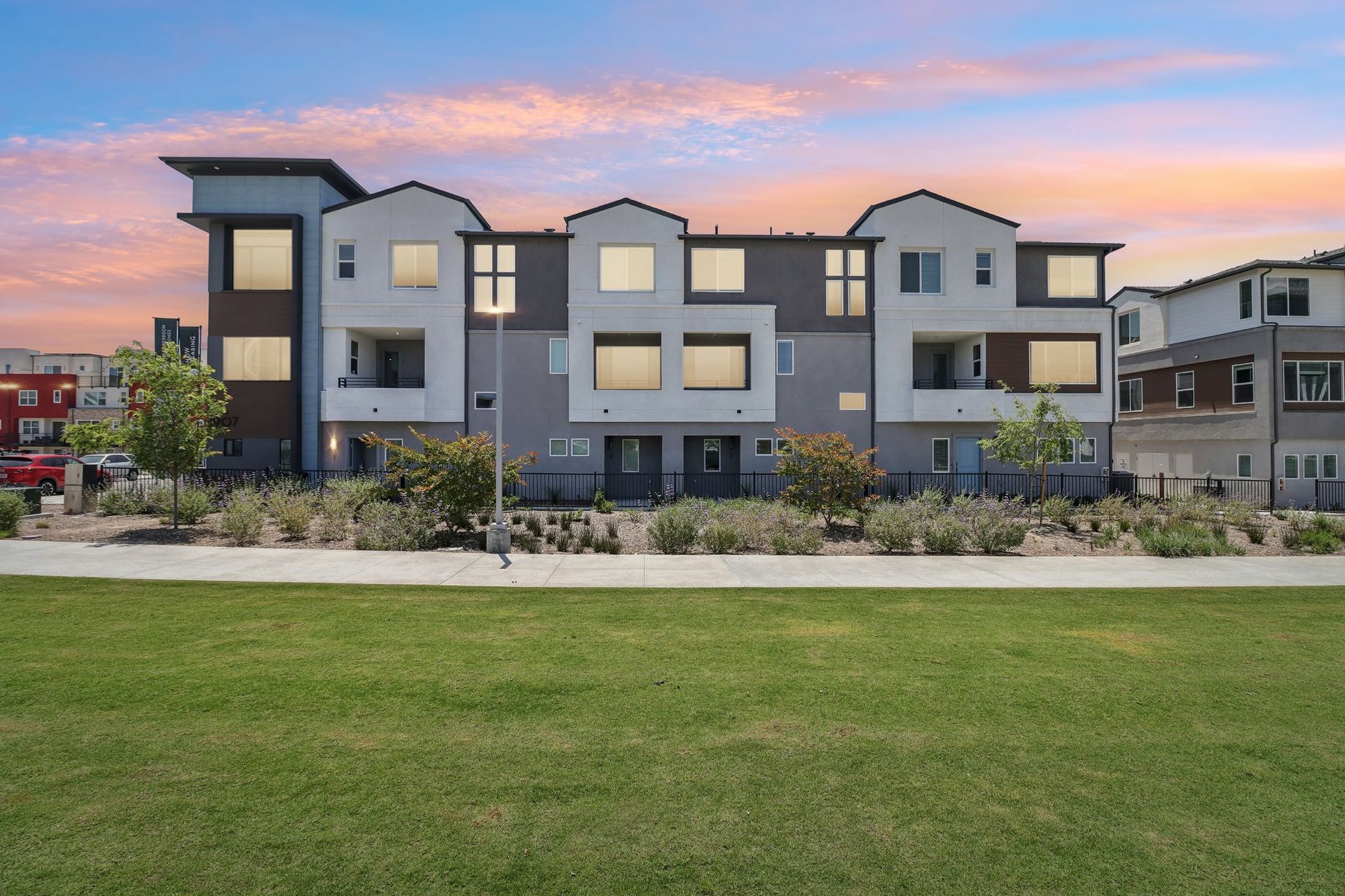 Townhomes with modern architecture against a colorful sunset sky and green lawn.