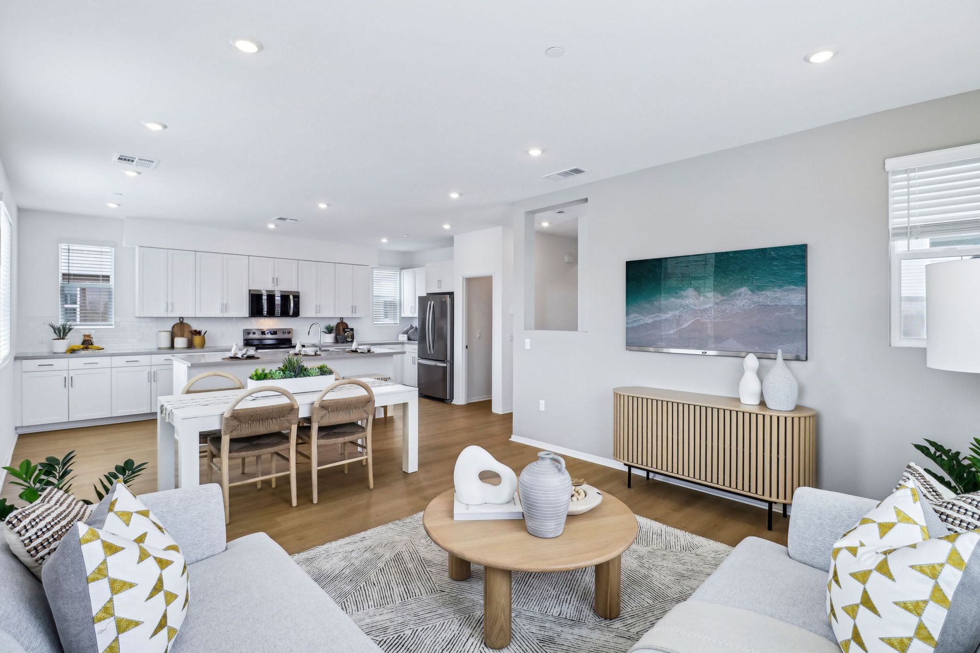 Open-concept living space: kitchen with white cabinets, dining, and living area with neutral tones, wood flooring, and art.