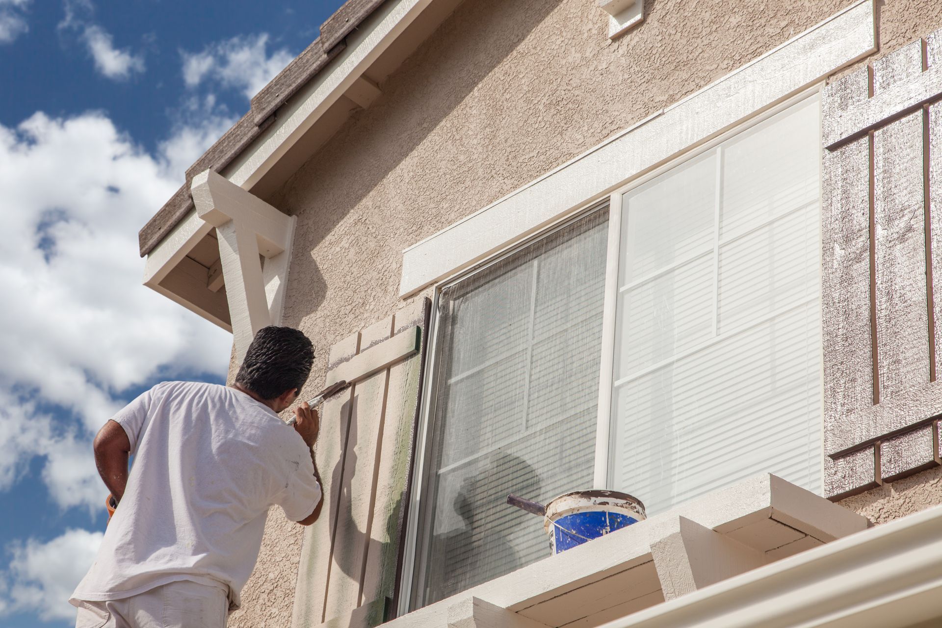Person painting an exterior wall beside a window under a blue sky