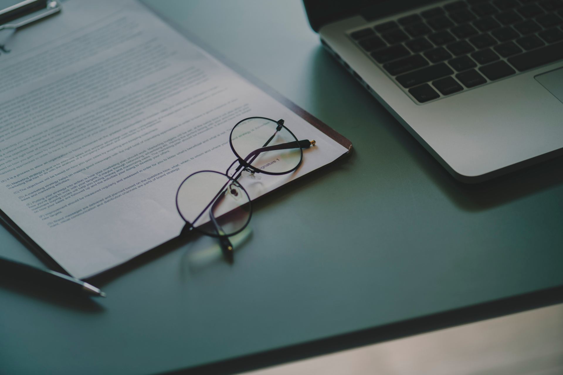 A pair of glasses is sitting on top of a clipboard next to a laptop.