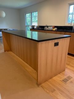 Light-wood kitchen island with a black countertop. A power outlet is visible. Light wood floor.