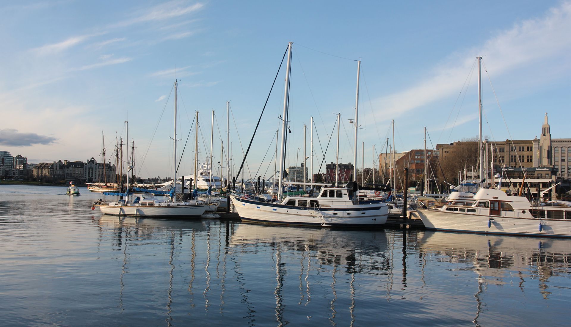 Boats docked in harbor, masts against blue sky, buildings in background reflecting in the water.