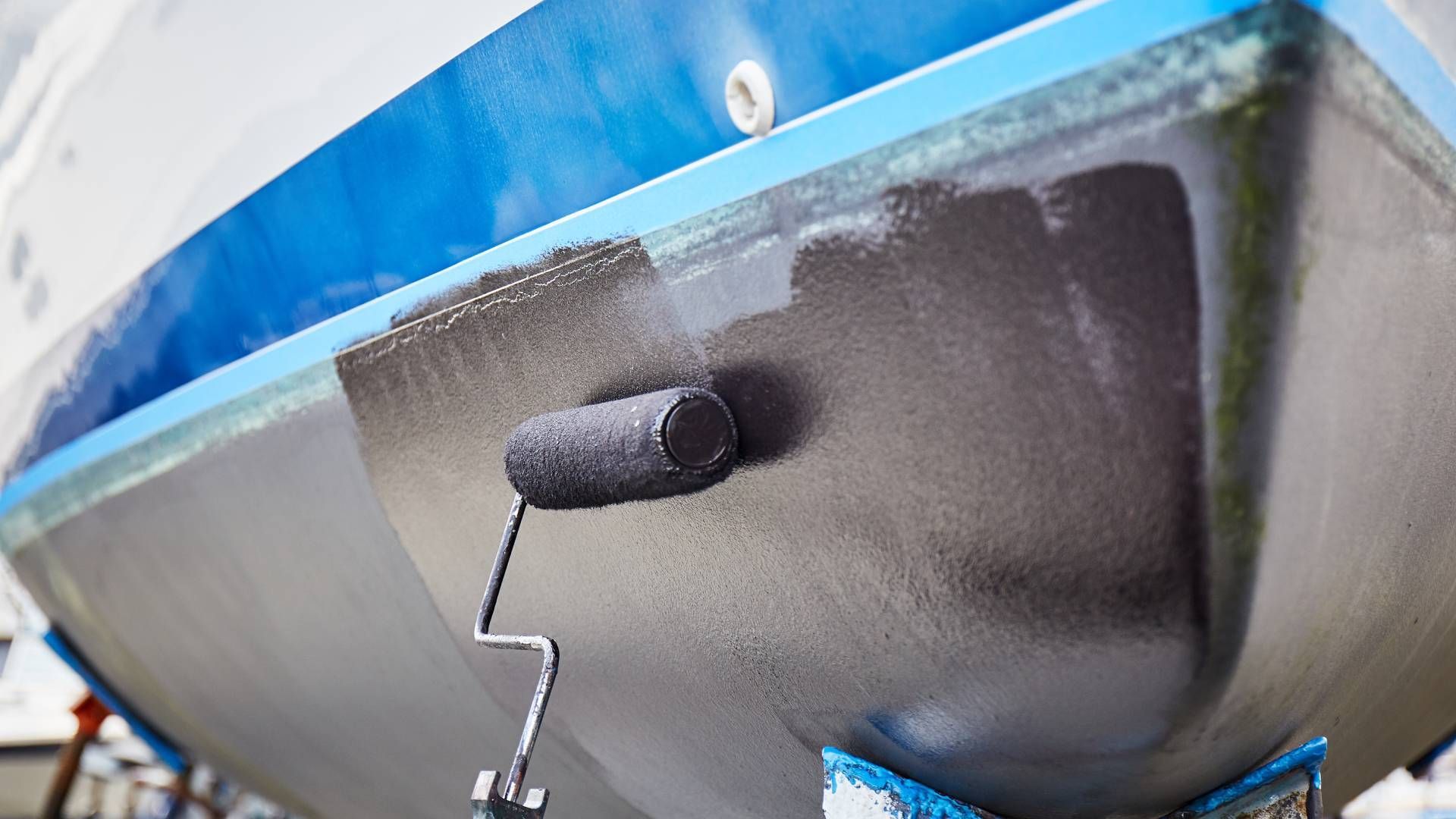 Boat hull being painted black with a roller. Blue and white boat in dry dock.